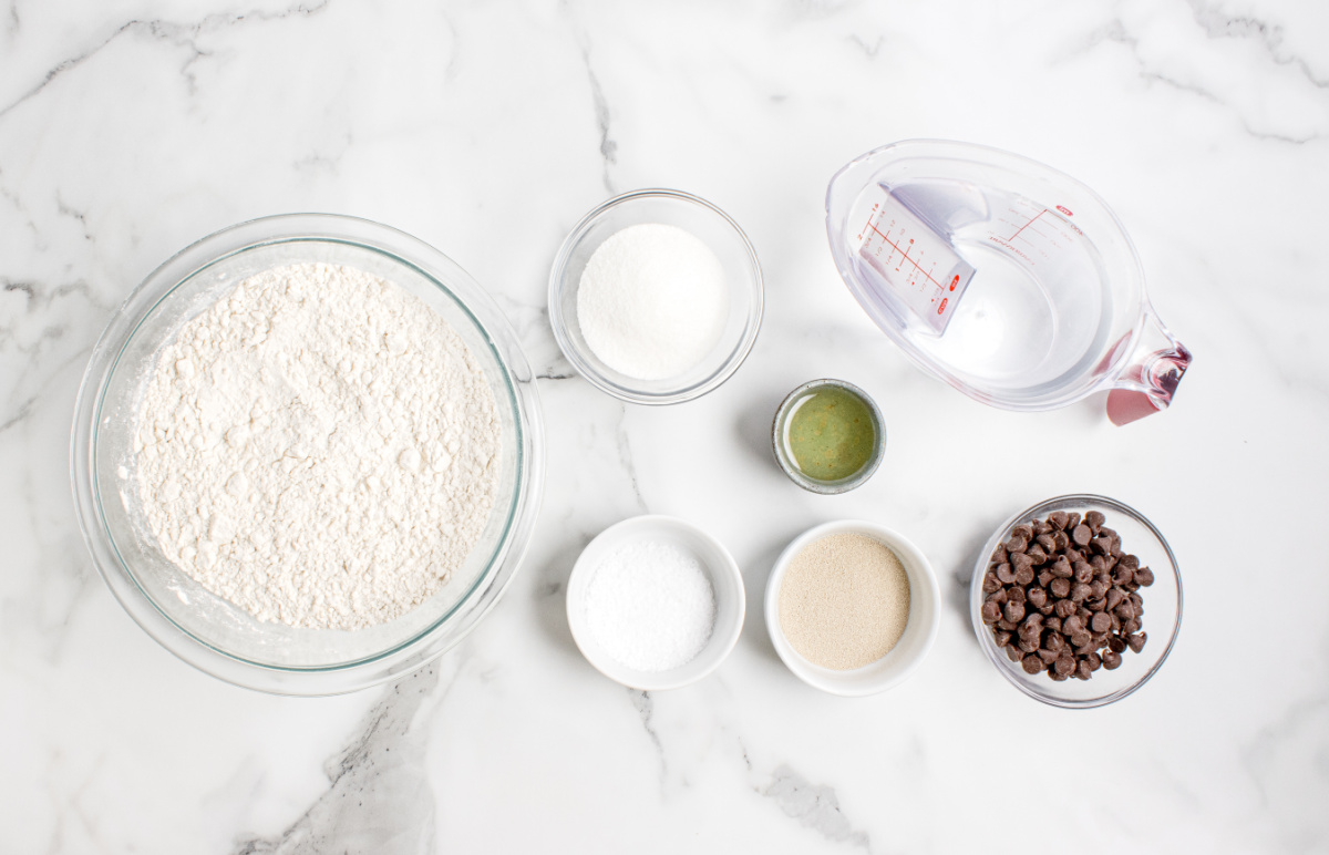 ingredients for chocolate chip bread including flour, yeast, sugar, salt, oil, water, and chocolate chips on a marble surface