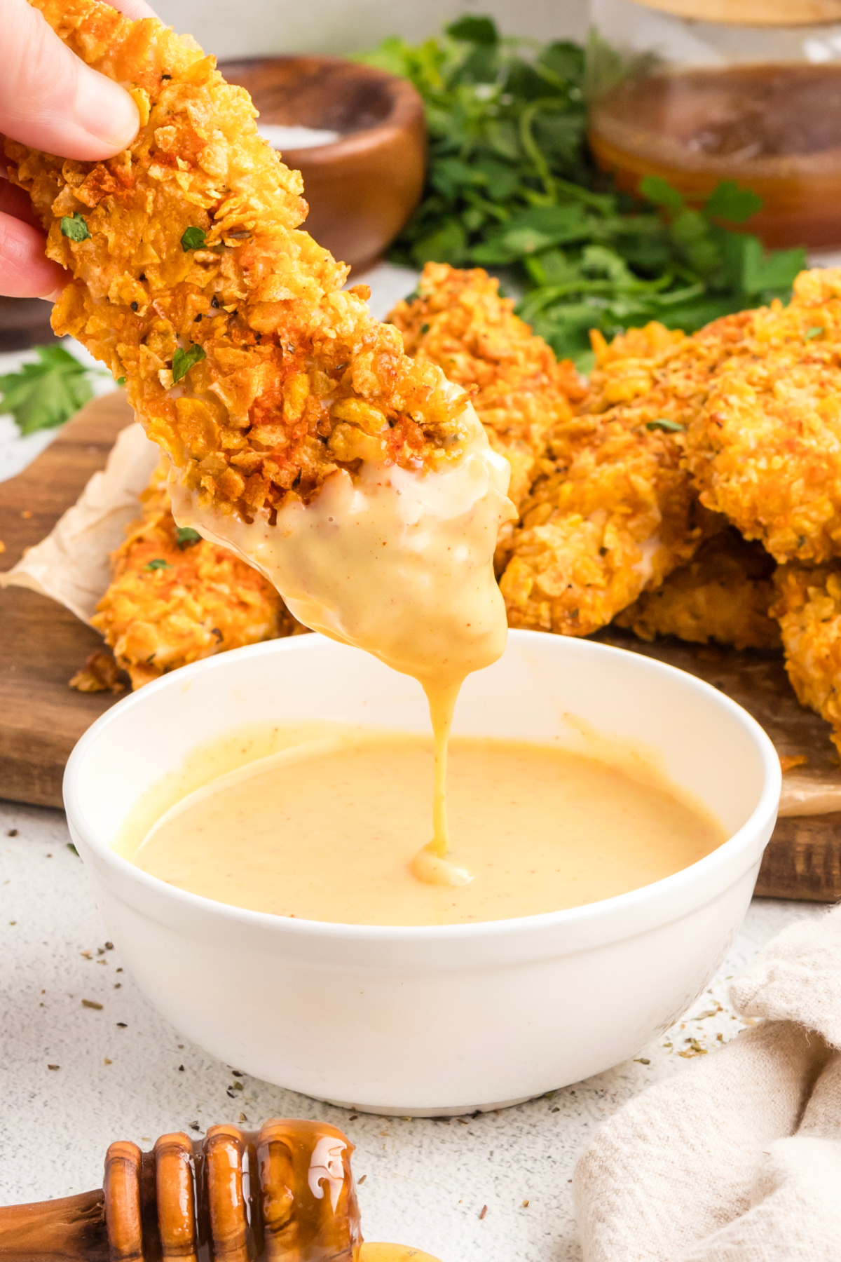 Hand holding crispy cornflake chicken tender dipped into homemade honey mustard sauce