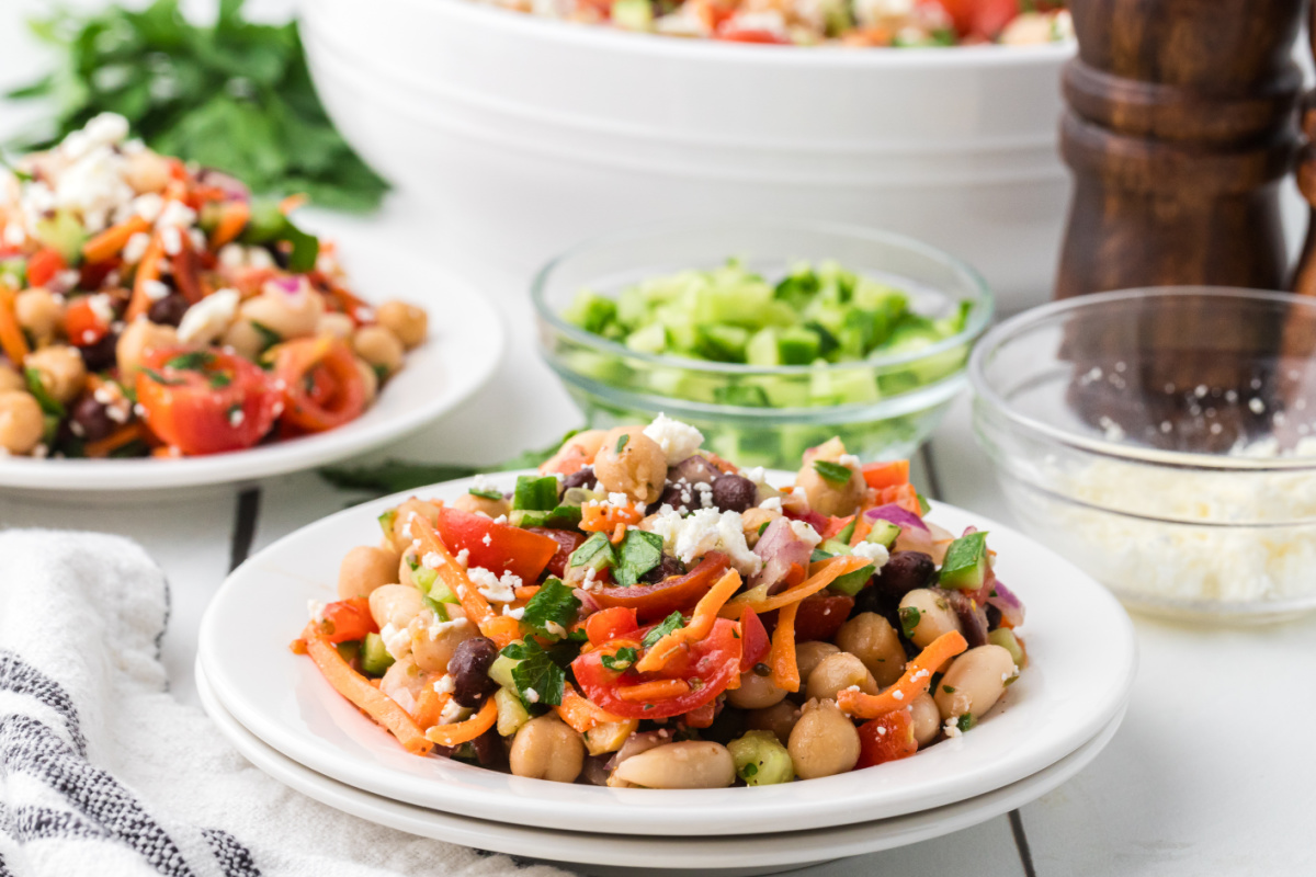 Dense bean salad recipe with chopped vegetables, feta, and three kinds of beans served cold for an easy make-ahead lunch. Dense bean salad recipe served on a white plate with feta cheese, chickpeas, black beans, tomatoes, cucumber, and parsley