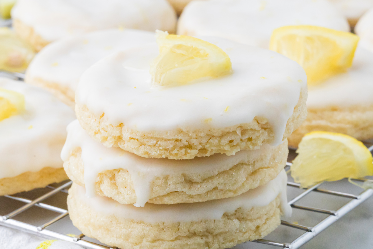 Glazed lemon cookies stacked on a cooling rack with thick lemon glaze and fresh lemon wedges