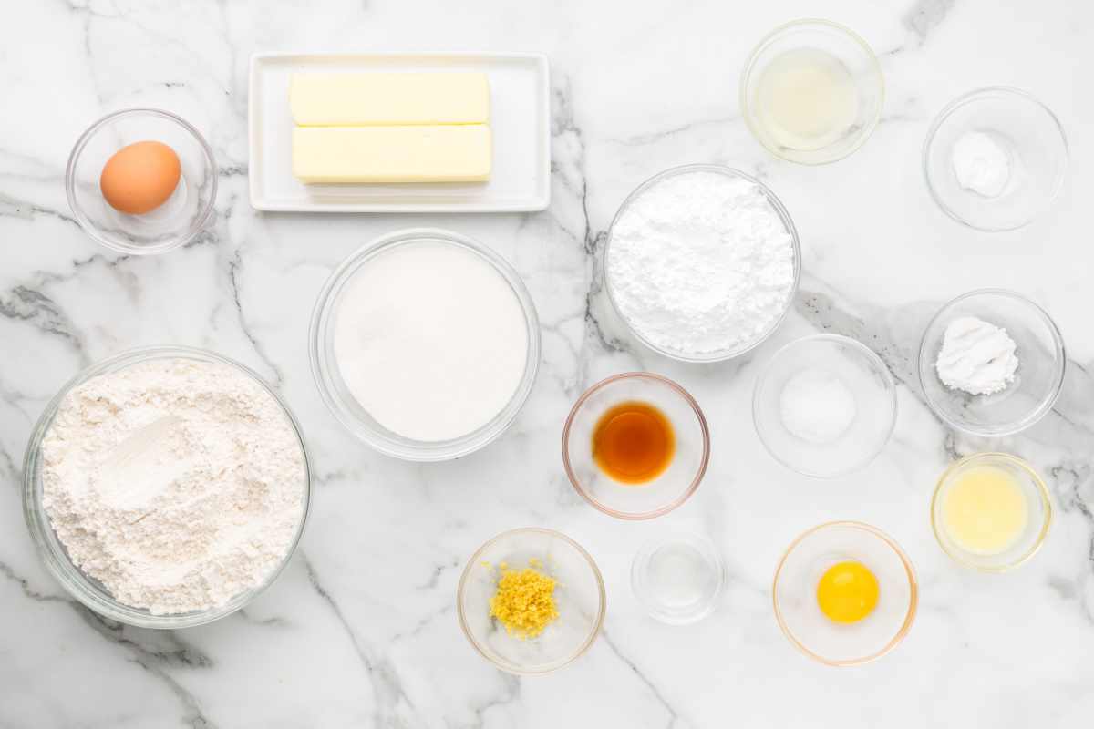 Ingredients for glazed lemon sugar cookies arranged on a marble surface including flour, butter, sugar, lemon zest, lemon juice, and eggs