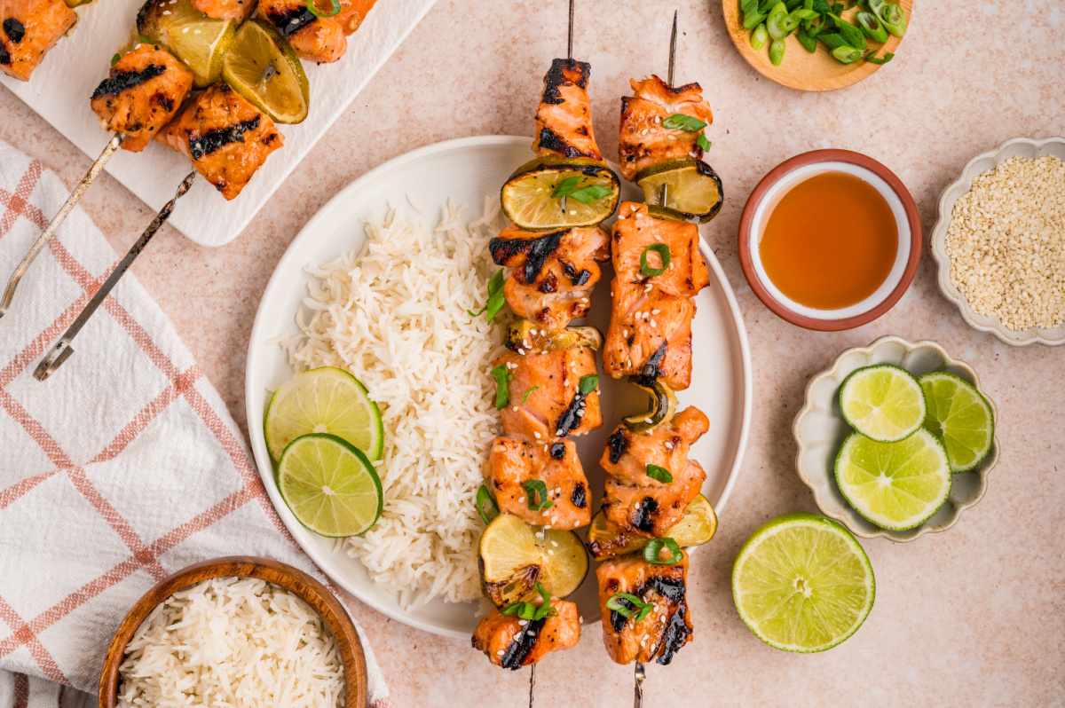 Overhead view of grilled honey soy lime salmon skewers served with white rice, grilled lime slices, sesame seeds, and green onions.