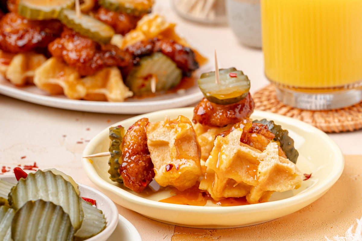 Hot Honey Peach Chicken and Waffle Bites on a Plate Hot honey peach chicken and waffle bites served on a plate with dill pickle chips and peach hot honey glaze.