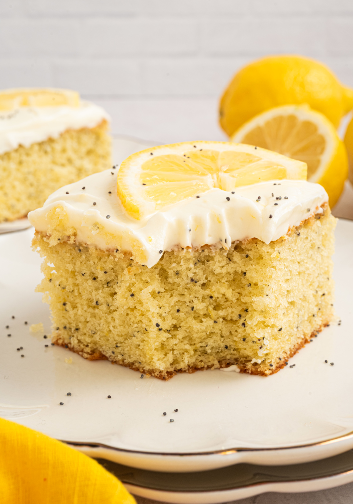 Lemon poppy seed cake slice with cream cheese frosting, topped with a fresh lemon slice and poppy seeds on a white plate