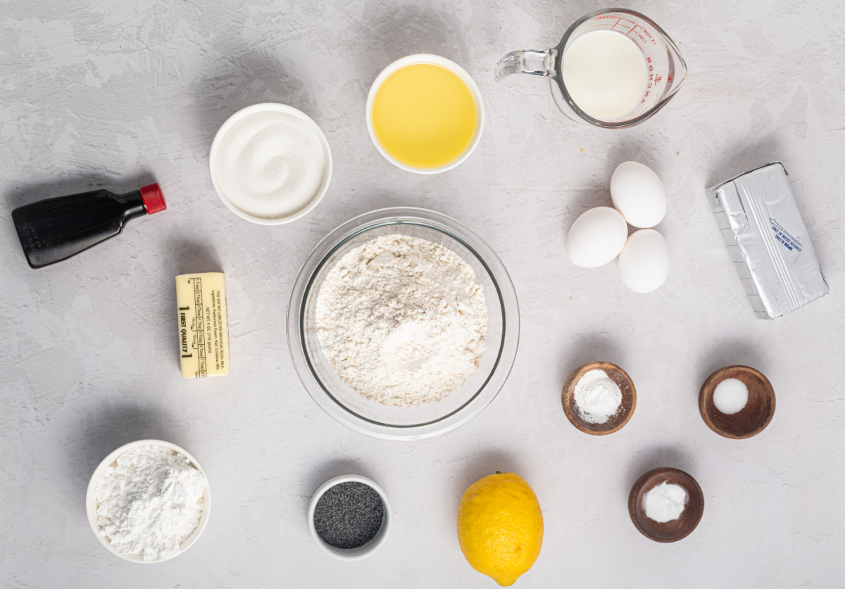 Ingredients for lemon poppy seed cake including flour, sugar, eggs, lemon, poppy seeds, milk, and butter arranged on a light surface