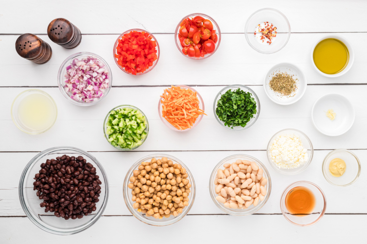 Ingredients for dense bean salad laid out with three kinds of beans, chopped vegetables, feta cheese, and lemon Dijon vinaigrette. Ingredients for dense bean salad including chickpeas, black beans, cannellini beans, feta, cucumber, tomatoes, carrots, and lemon Dijon dressing