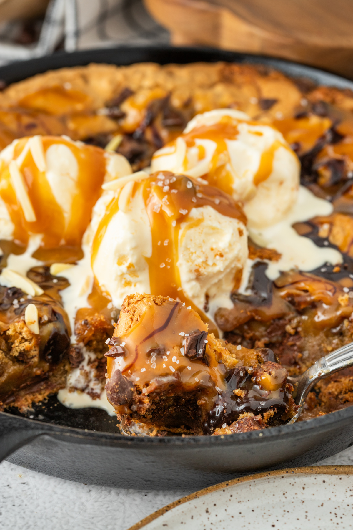 Spoon lifting a gooey bite of salted caramel chocolate chip skillet cookie with molten caramel, melted chocolate, vanilla ice cream, and flaky sea salt in a cast iron skillet.