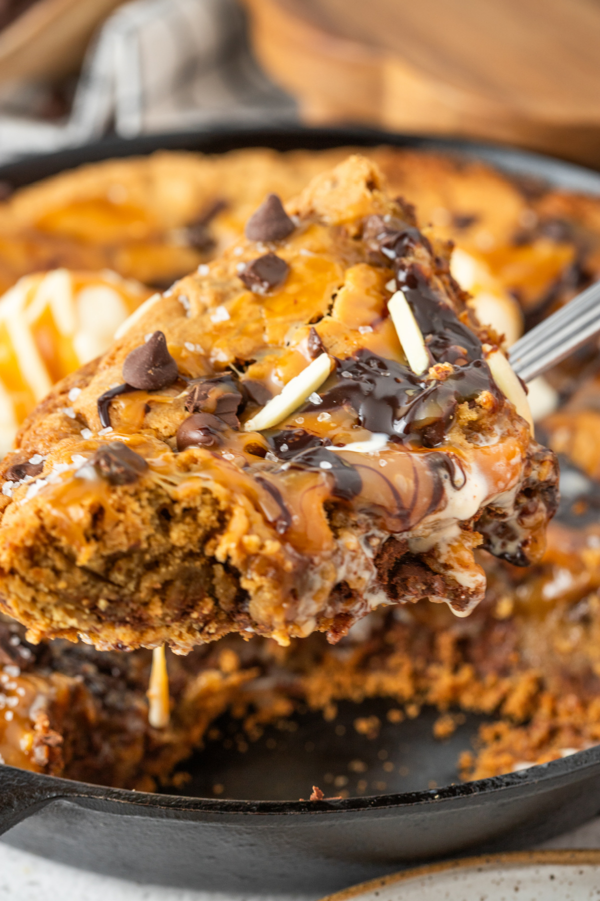 Slice of salted caramel cookie skillet lifted from a cast iron pan with melted chocolate, caramel drizzle, flaky sea salt, and melting vanilla ice cream.