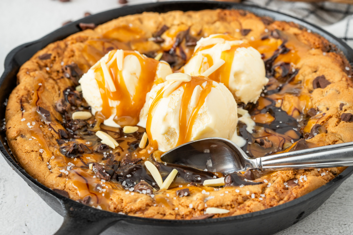 Warm salted caramel skillet cookie topped with vanilla ice cream, chocolate drizzle, flaky sea salt, and molten caramel in a cast iron skillet.