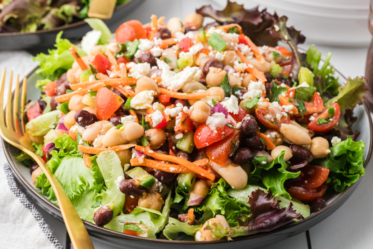 Three bean salad with feta cheese and chopped vegetables served over lettuce with lemon Dijon dressing. Three bean salad with feta cheese, chickpeas, black beans, cannellini beans, tomatoes, carrots, and cucumber over lettuce greens