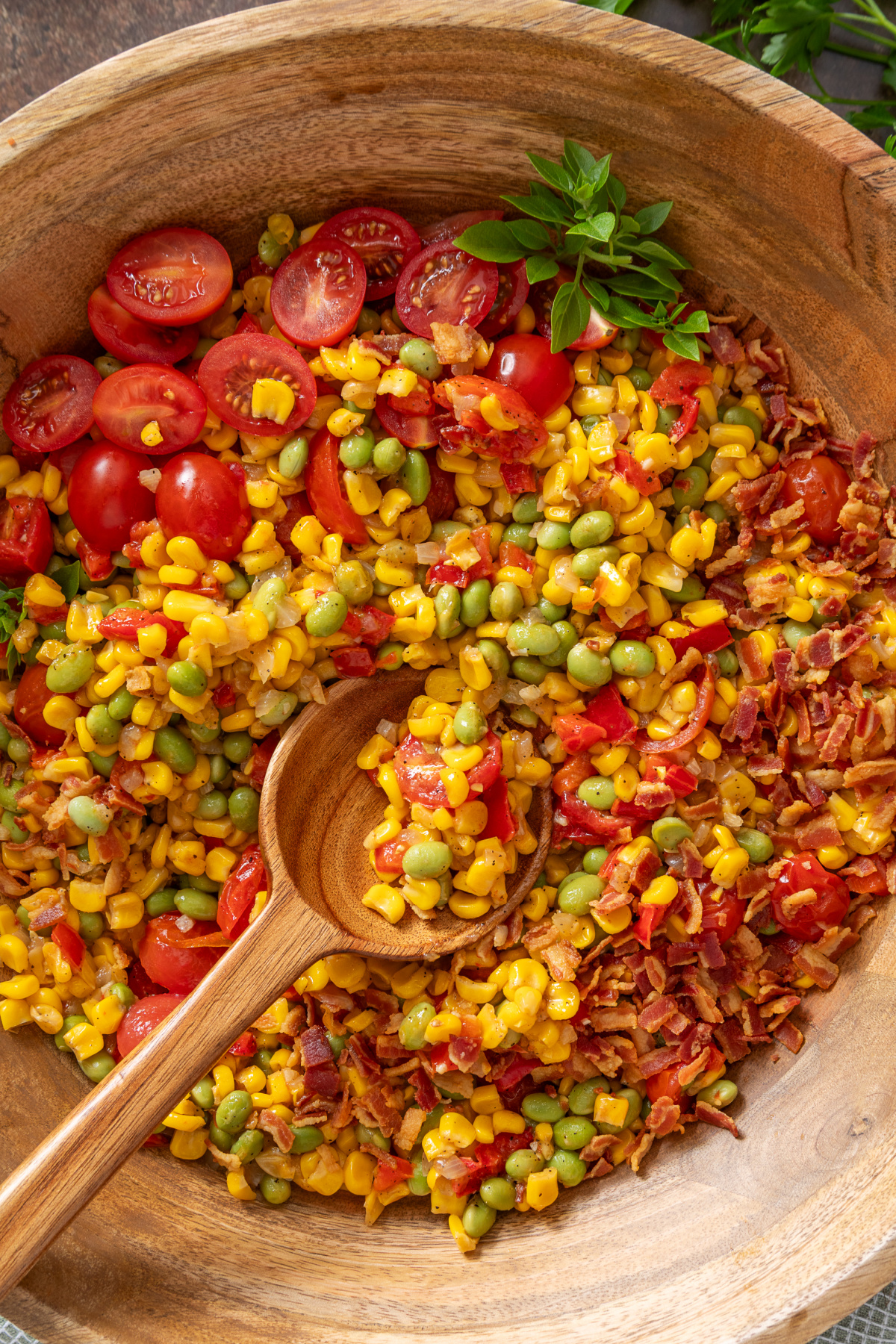 Overhead view of a bacon and corn side dish with edamame, tomatoes, peppers, and onions in a wooden bowl
