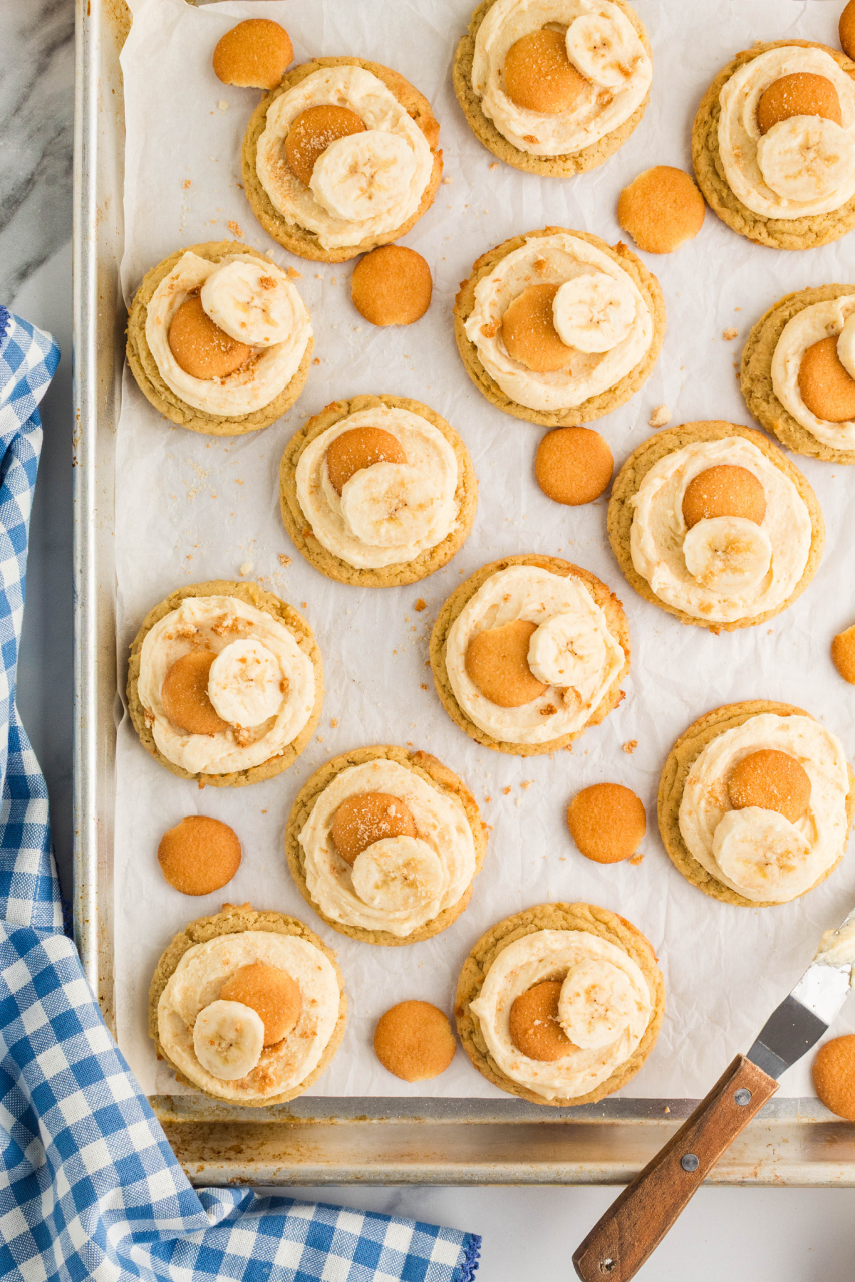 Banana pudding cookies arranged on a parchment-lined baking sheet with brown butter cream cheese frosting, mini vanilla wafers, and fresh banana slices.