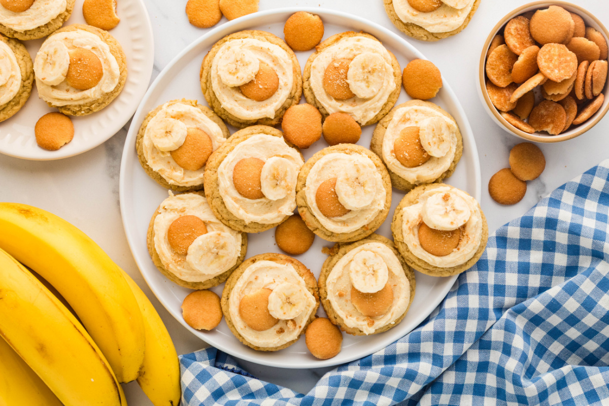 Platter of banana pudding cookies frosted with brown butter cream cheese frosting and topped with vanilla wafers and fresh banana slices.