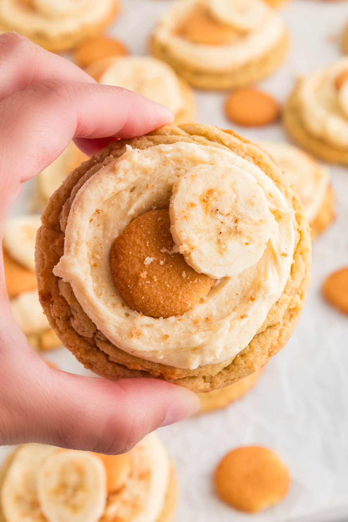 Hand holding a frosted banana pudding cookie topped with a mini vanilla wafer and fresh banana slice.