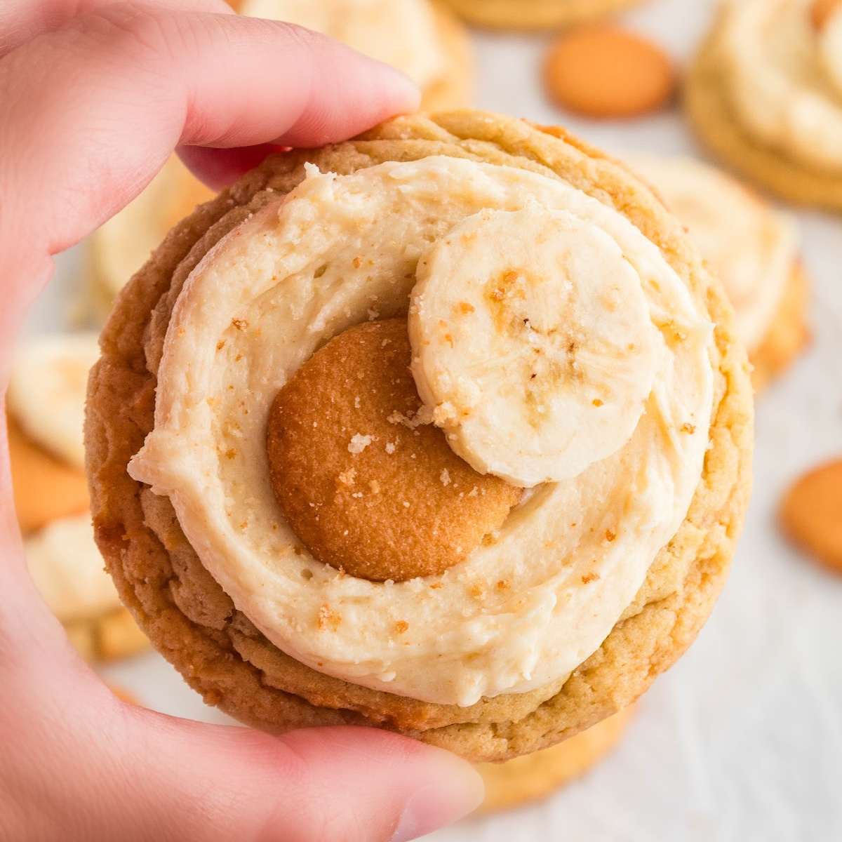 Hand holding a banana pudding cookie topped with brown butter cream cheese frosting, a mini vanilla wafer, and a fresh banana slice.