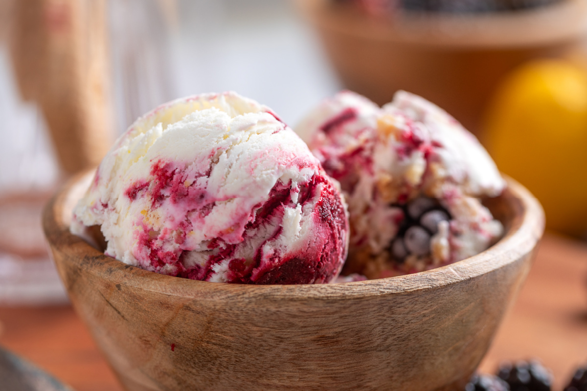 Blackberry Crumble à la Mode Ice Cream has thick blackberry swirls in every scoop with buttery shortbread crumble throughout. Two scoops of Blackberry Crumble à la Mode Ice Cream with thick blackberry ribbons in a wooden bowl.