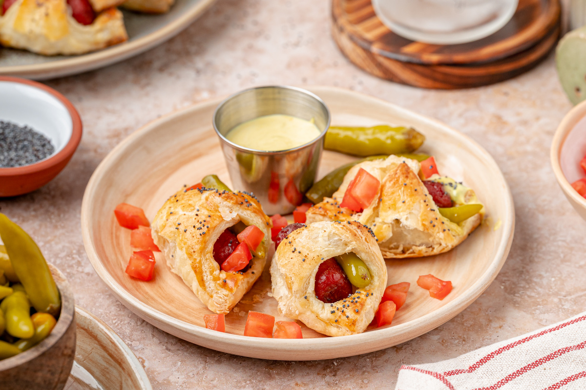 Chicago hot dog pigs in a blanket baked with sport peppers and served with a creamy mustard relish dipping sauce. Close-up of Chicago hot dog pigs in a blanket with sport peppers, diced tomato, and creamy mustard relish dip.