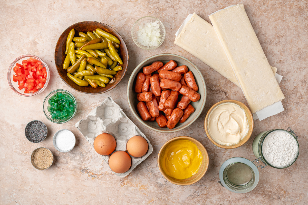 Everything needed to make Chicago hot dog pigs in a blanket, including sport peppers, beef cocktail franks, puff pastry, and mustard relish dip ingredients. Ingredients for Chicago hot dog pigs in a blanket including puff pastry, beef cocktail franks, sport peppers, mustard, mayonnaise, relish, onion, tomato, and poppy seeds.