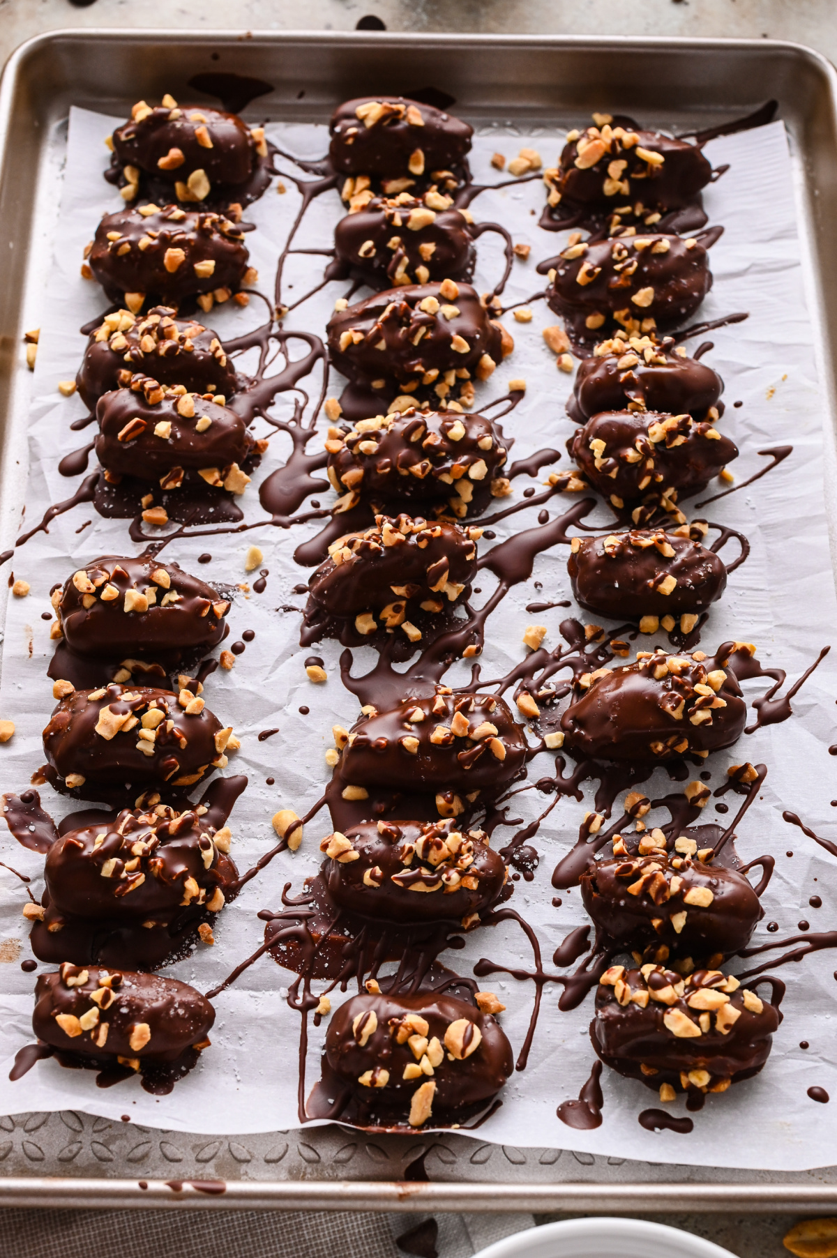 Freshly made stuffed dates setting on a parchment-lined tray Chocolate-covered stuffed dates topped with chopped peanuts and flaky sea salt on a parchment-lined tray