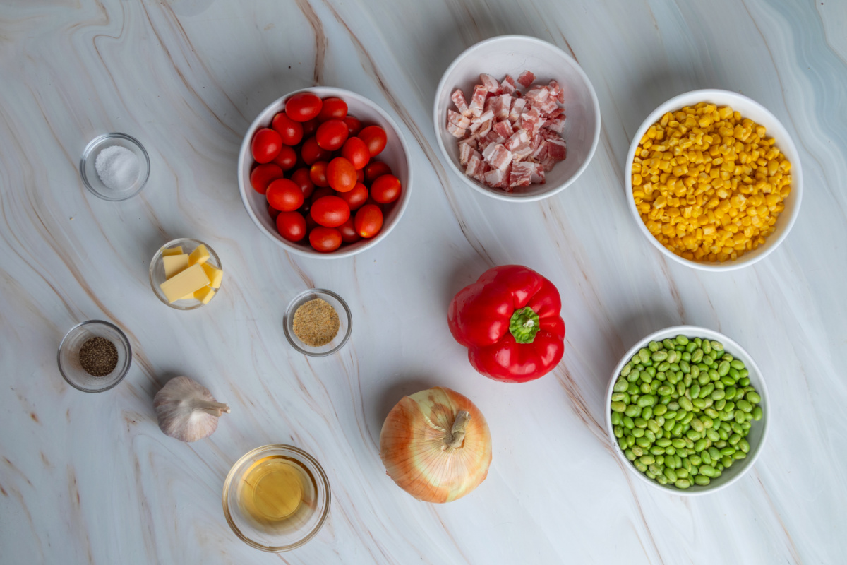 Ingredients for corn succotash including sweet corn, edamame, bacon, tomatoes, bell pepper, onion, garlic, butter, and apple cider vinegar