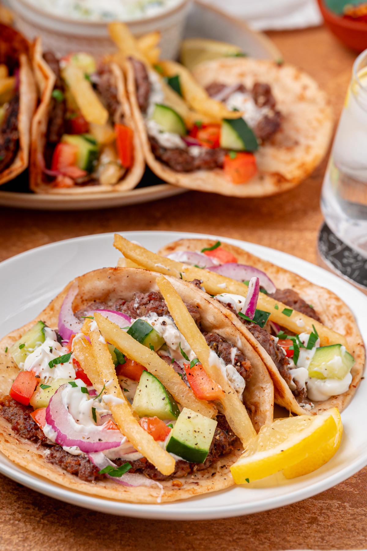 Greek-inspired smashed tacos served with tzatziki, fresh vegetables, fries, and lemon wedges. Greek-inspired smashed tacos topped with seasoned beef, tzatziki, fries, cucumber, tomato, and red onion on a white plate.