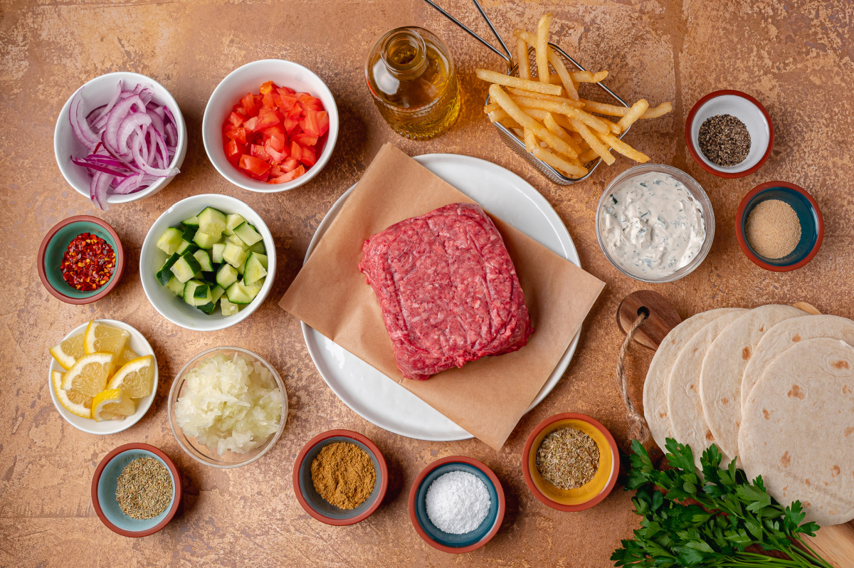 Everything needed to make gyro smashed tacos, from the seasoned beef to the tzatziki and fries. Ingredients for gyro smashed tacos including ground beef, tortillas, tzatziki, fries, cucumber, tomato, red onion, parsley, and seasonings.