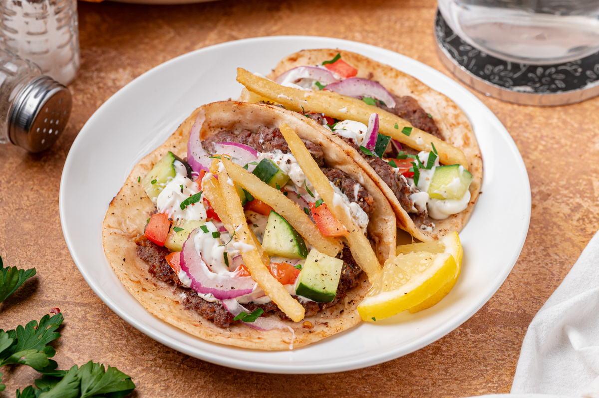 Gyro smashed tacos served with fresh vegetables, tzatziki, fries, and lemon. Gyro smashed tacos topped with tzatziki, cucumber, tomato, red onion, and fries on a white plate with lemon wedges.