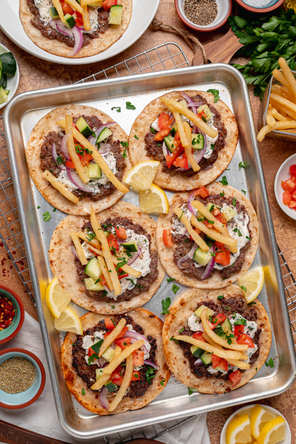 A sheet pan full of gyro smashed tacos ready for serving with classic gyro toppings. Gyro smashed tacos arranged on a sheet pan with tzatziki, cucumber, tomato, red onion, fries, and lemon wedges.