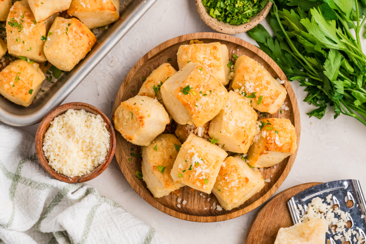 A close-up of homemade Parmesan bread bites baked until golden and finished with Parmesan cheese Parmesan bread bites on a wooden platter topped with Parmesan cheese and fresh parsley