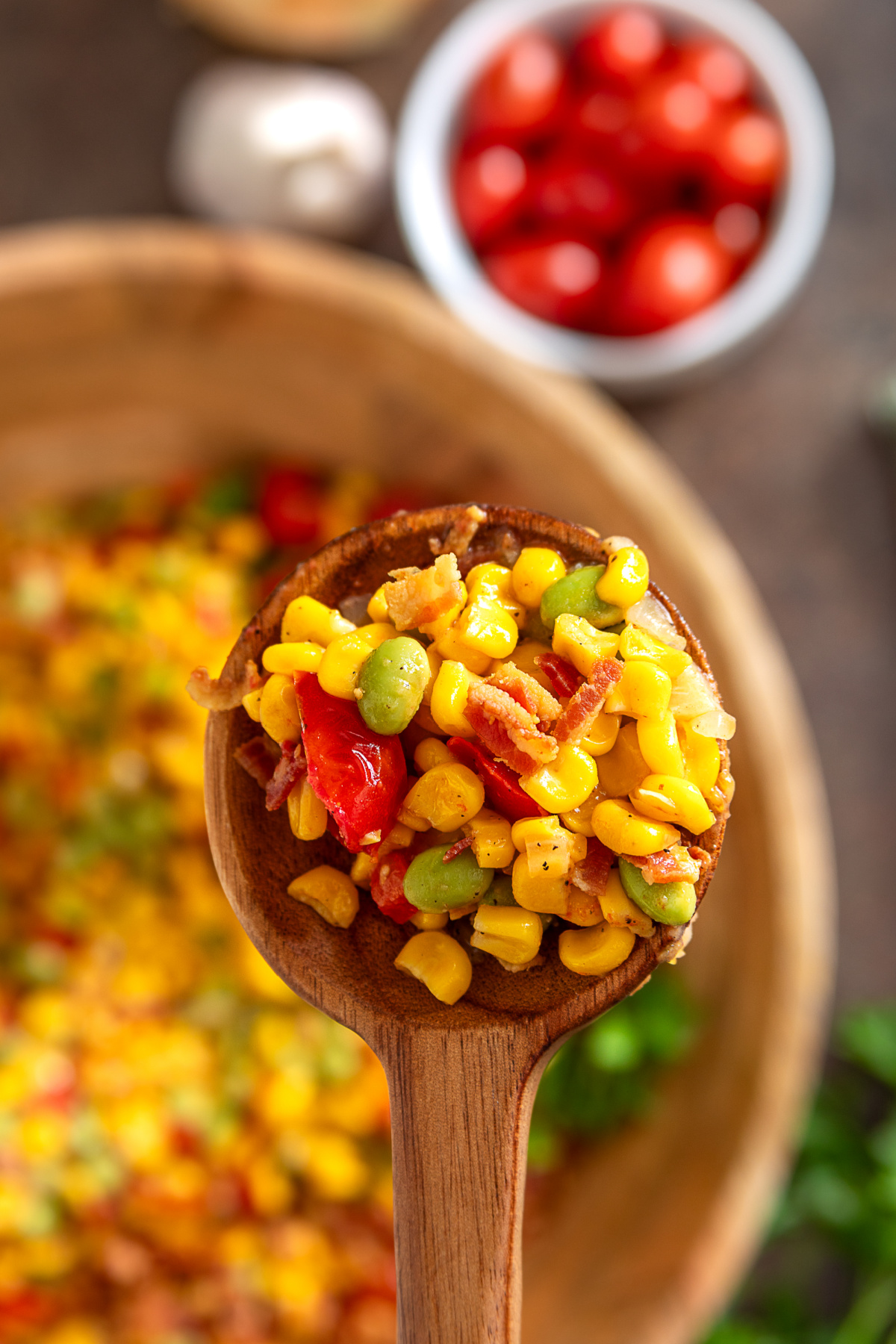 Wooden spoon holding sweet corn, edamame, bacon, peppers, and tomatoes from a summer corn side dish