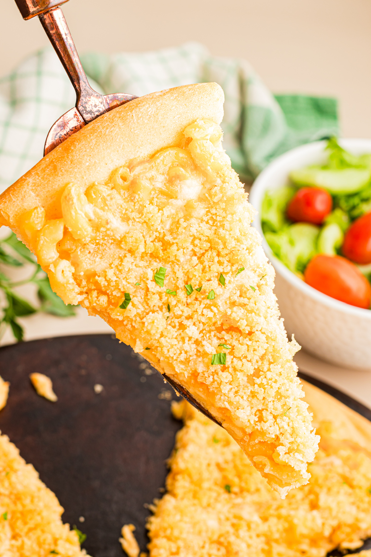 Baked mac and cheese pizza slice lifted on a pizza server with a golden buttery breadcrumb topping and thick homemade macaroni and cheese.