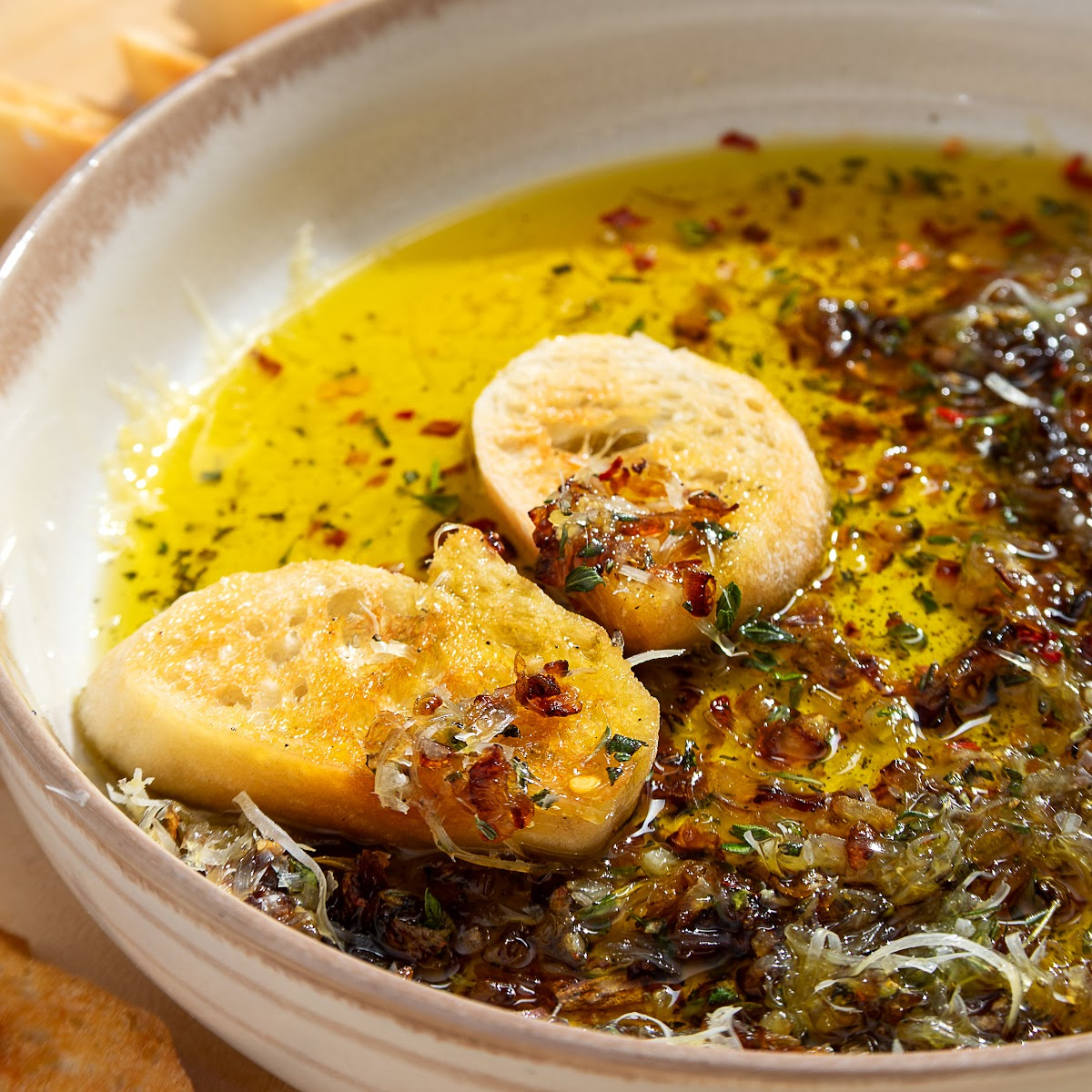 Toasted bread resting in bread dipping oil with caramelized shallots, garlic, fresh herbs, Parmesan, and olive oil.