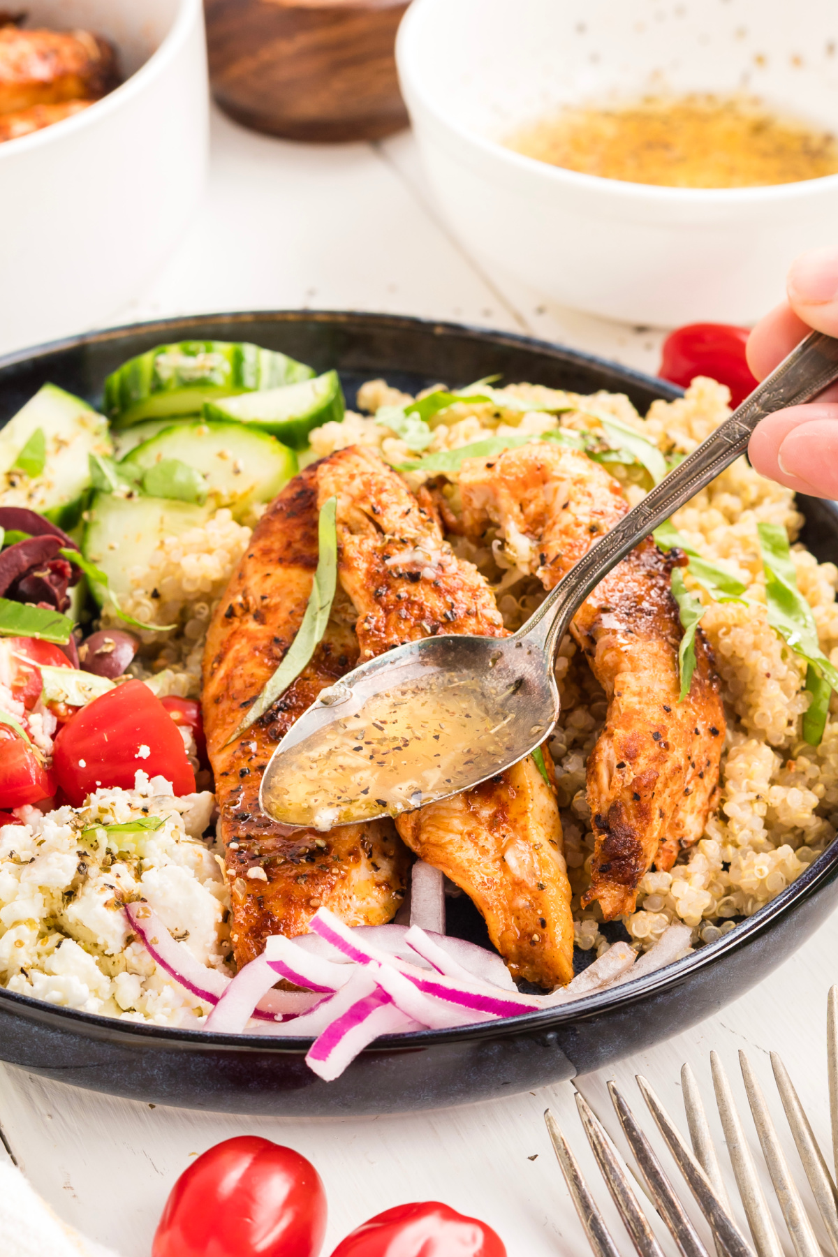 Drizzling Lemon Vinaigrette Over Mediterranean Chicken Bowl Spoon drizzling homemade lemon vinaigrette over a Mediterranean chicken bowl with grilled chicken, quinoa, feta, tomatoes, cucumber, olives, and red onion.