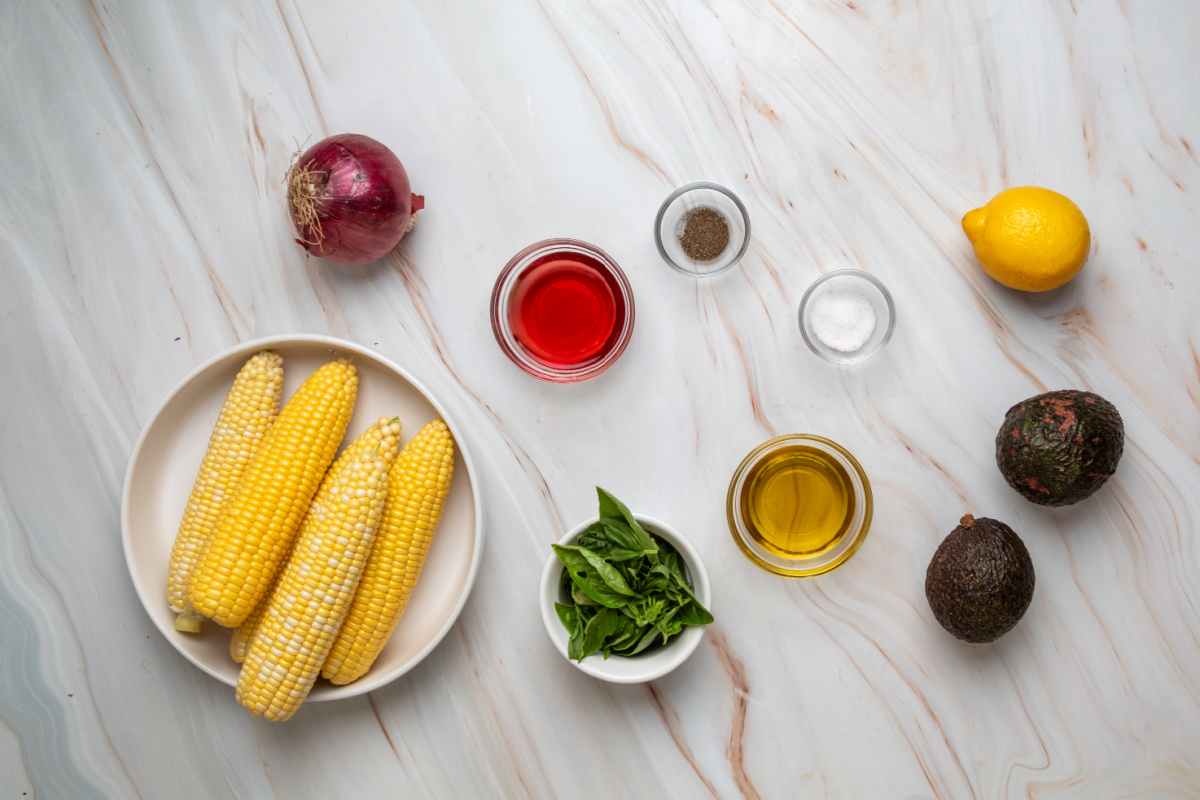 Ingredients for grilled corn salad with avocado and basil, including fresh corn, avocado, basil, red onion, olive oil, lemon, and red wine vinegar.