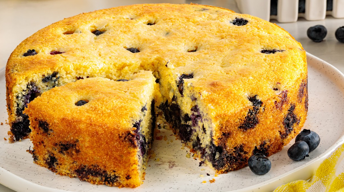 A slice reveals the tender crumb and fresh blueberries throughout this lemon blueberry cornbread. Lemon blueberry cornbread with one slice removed, showing the golden crust and blueberries throughout the tender crumb.