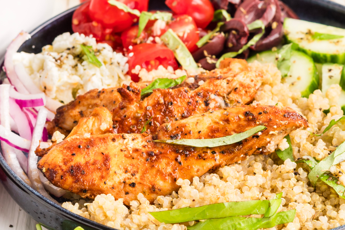 Close-up of a Mediterranean chicken bowl with grilled chicken, quinoa, feta, tomatoes, cucumber, Kalamata olives, and red onion.
