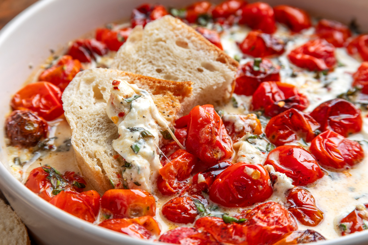Serving Roasted Tomato Burrata Dip with crusty bread. Crusty bread scooping creamy burrata with roasted grape tomatoes, basil, garlic, and roasted tomato juices.