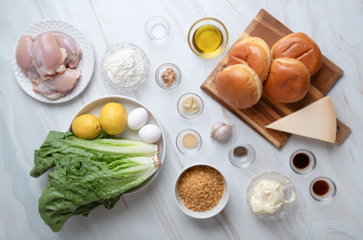 Chicken Caesar salad sandwich ingredients Ingredients for a chicken Caesar salad sandwich, including chicken thighs, romaine lettuce, Parmesan cheese, potato chips, sandwich rolls, eggs, lemons, olive oil, and Caesar dressing ingredients.