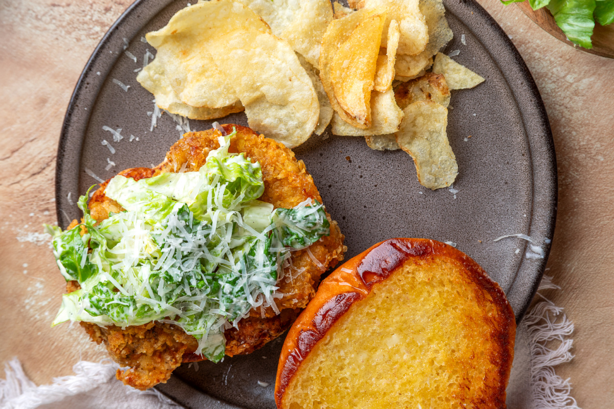 Chicken Caesar salad sandwich overhead view Overhead view of a chicken Caesar salad sandwich with crispy potato chip-crusted chicken, homemade Caesar salad, toasted brioche bun, and potato chips.