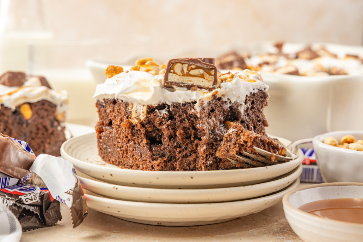 Chocolate caramel poke cake with pudding filling, salted peanuts and Snickers candy on top Chocolate cake slice showing pudding and caramel filling beneath whipped topping and chopped Snickers