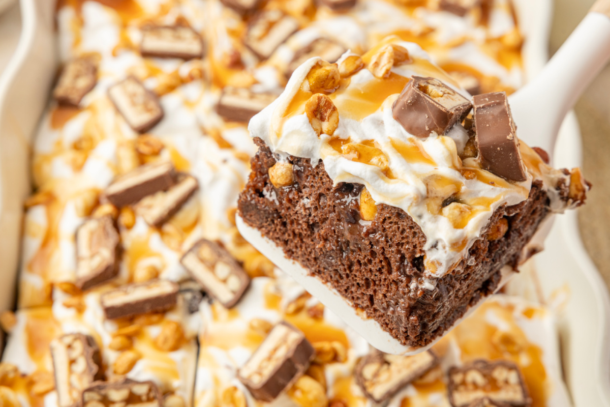 Slice of chocolate pudding poke cake topped with whipped topping, caramel, peanuts and chopped Snickers bars Chocolate poke cake slice showing pudding and caramel filling with roasted salted peanuts