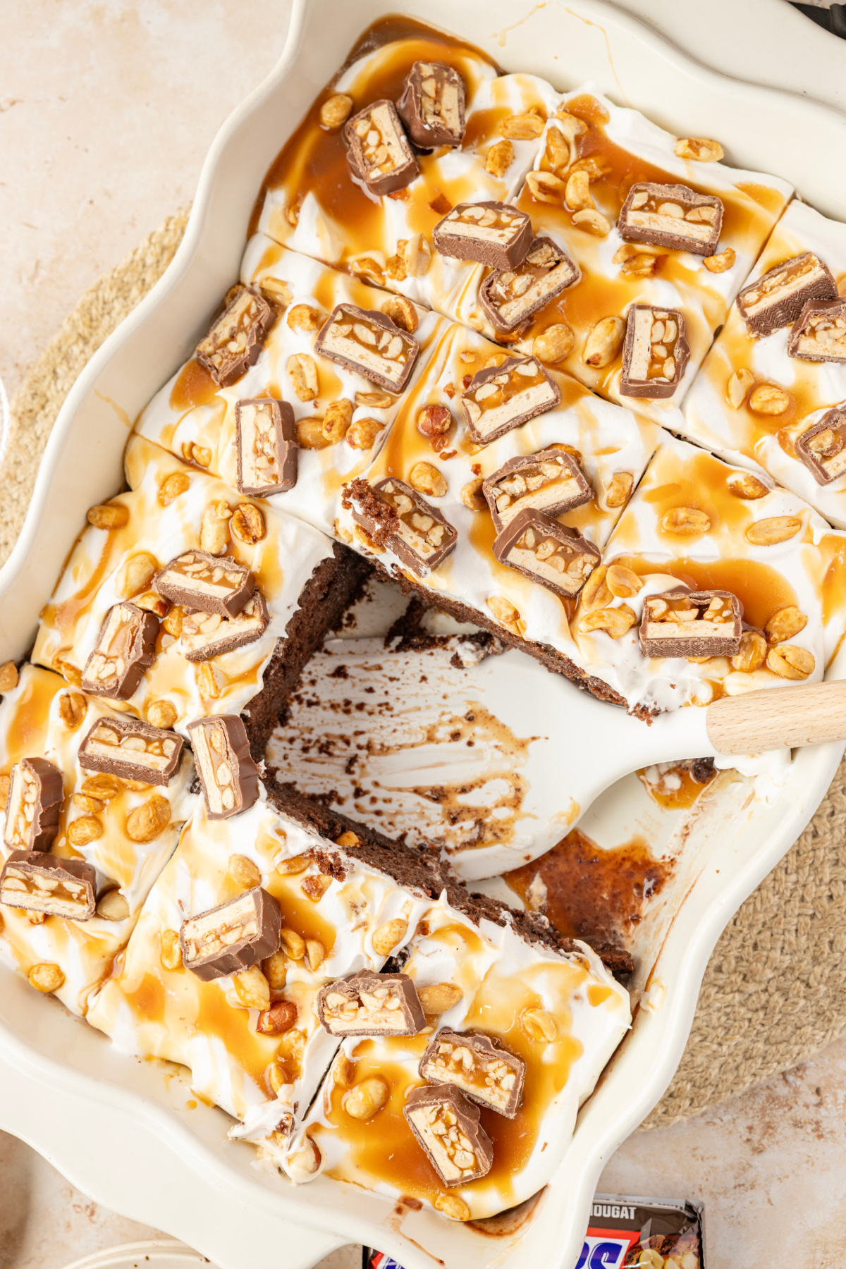 Sliced chocolate poke cake with pudding filling, whipped topping, caramel, peanuts and Snickers candy Chocolate pudding poke cake cut into squares with caramel, salted peanuts and chopped Snickers bars