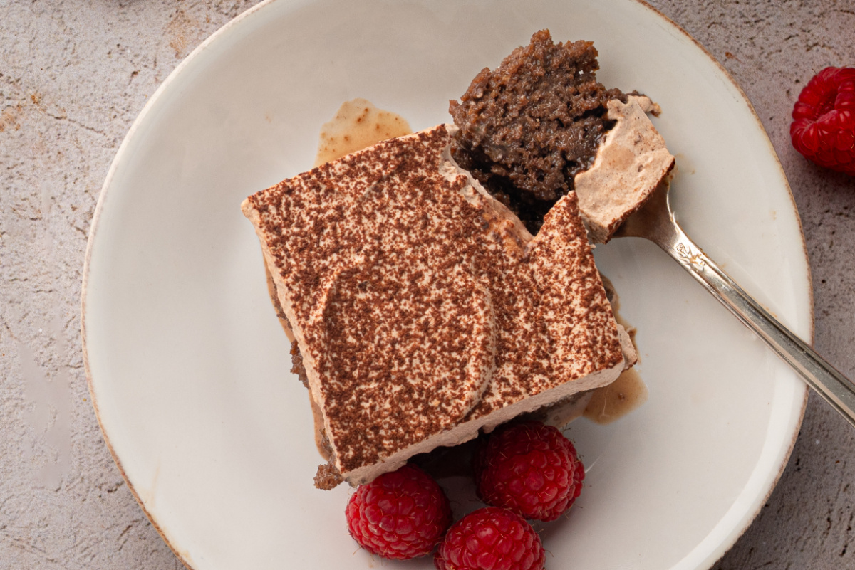 Chocolate tres leches cake topped with cocoa-dusted whipped cream and served with raspberries. Chocolate tres leches cake with a bite removed, showing the milk-soaked chocolate cake beneath chocolate whipped cream.