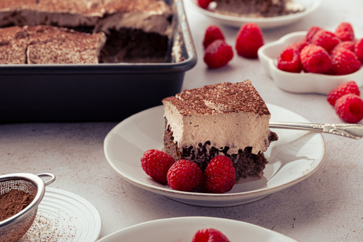 Chocolate tres leches cake served with fresh raspberries and cocoa-dusted whipped cream. Slice of chocolate tres leches cake with chocolate whipped cream and raspberries beside the baking pan.