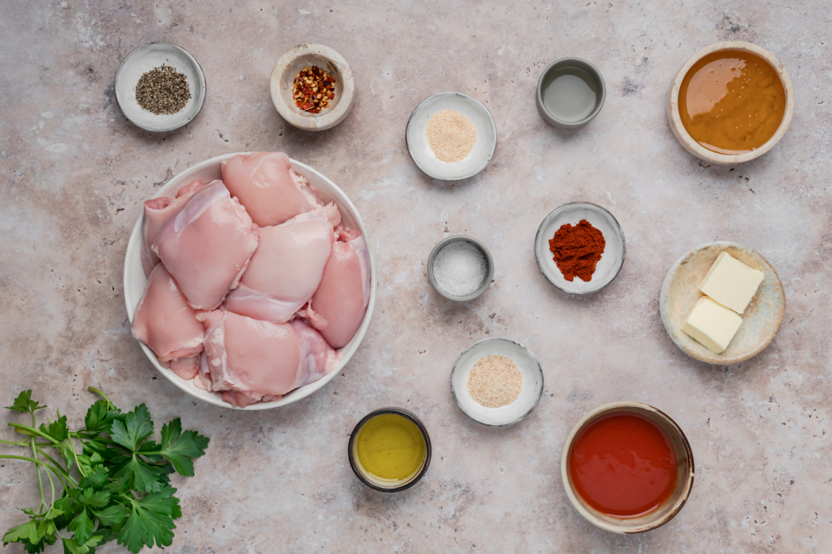 Ingredients for hot honey chicken thighs, including boneless chicken thighs, honey, hot sauce, butter, spices, olive oil, and parsley.