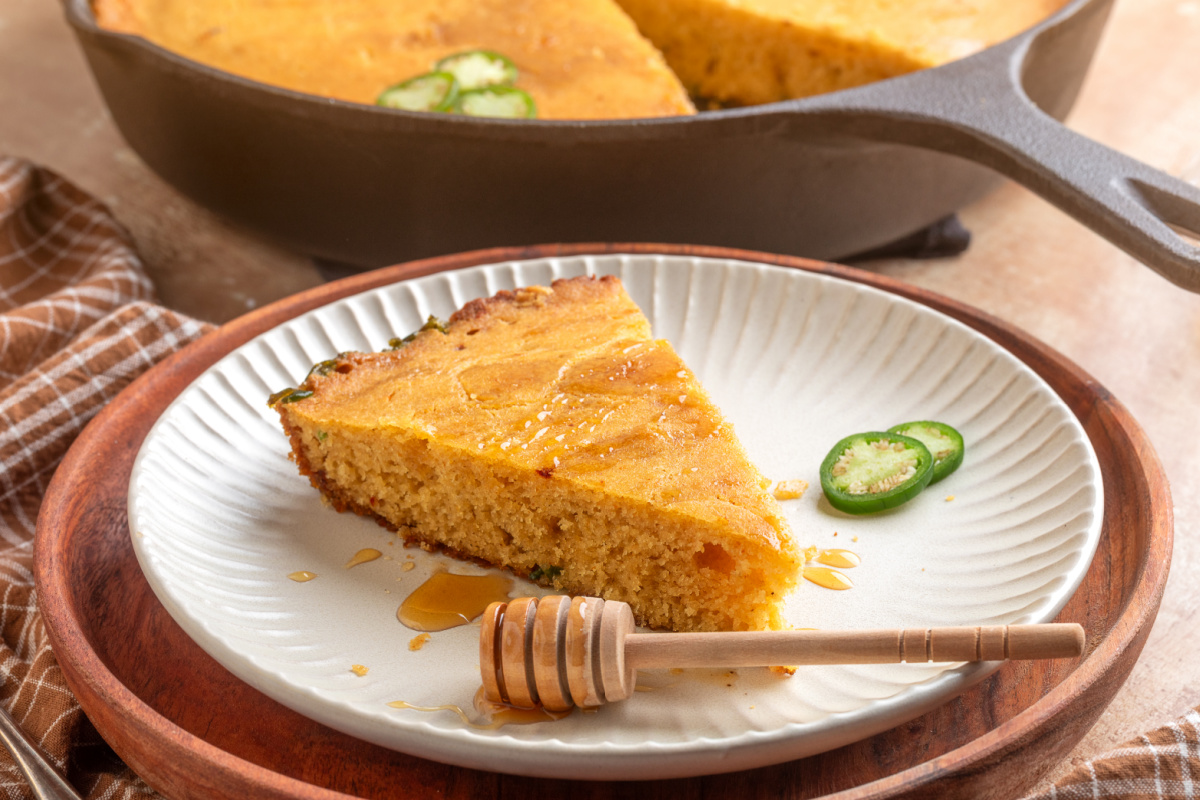 Chipotle cornbread with fresh jalapeños and hot honey served warm from the skillet Slice of hot honey jalapeño cornbread served with the cast-iron skillet in the background