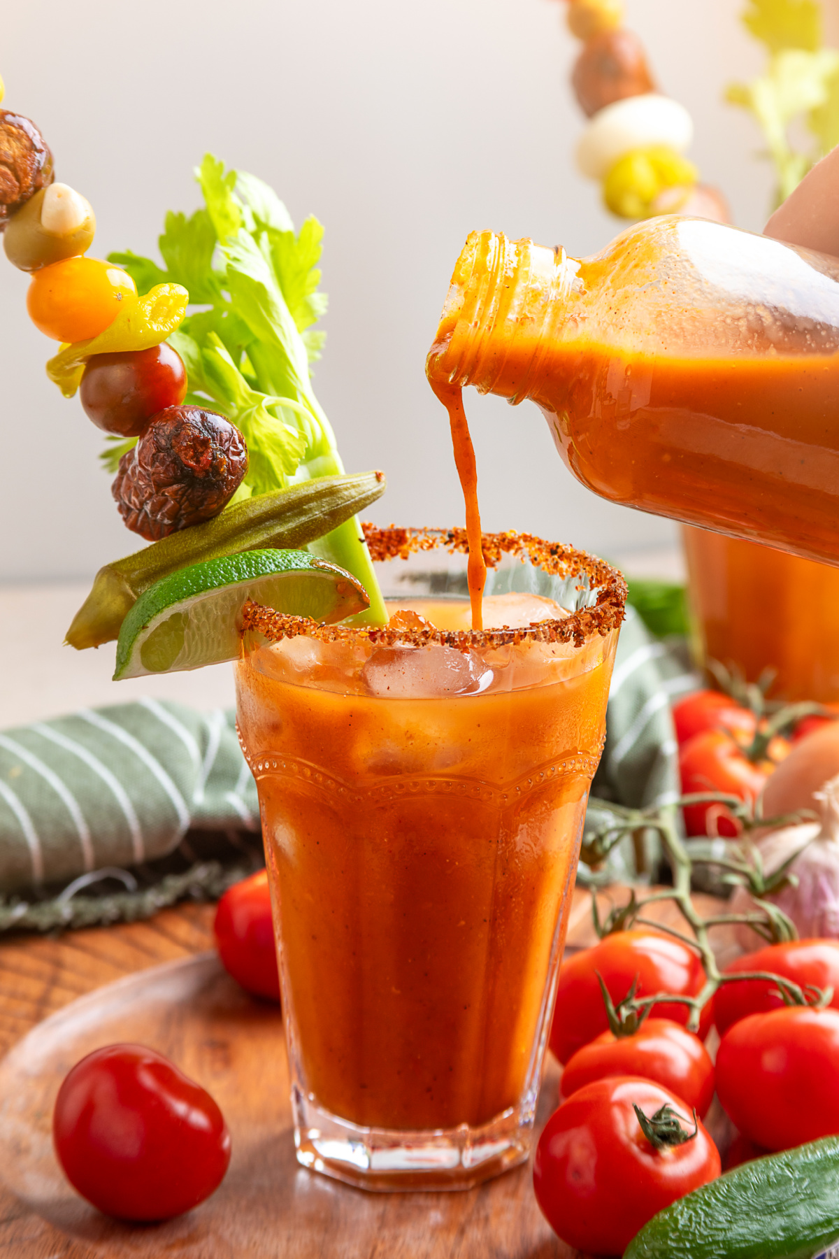 Homemade smoked tomato Bloody Mary mix poured over ice with celery, lime, pickled okra and smoked sausage Smoked tomato Bloody Mary mix being poured into an ice-filled glass with a Tajín rim and garnishes