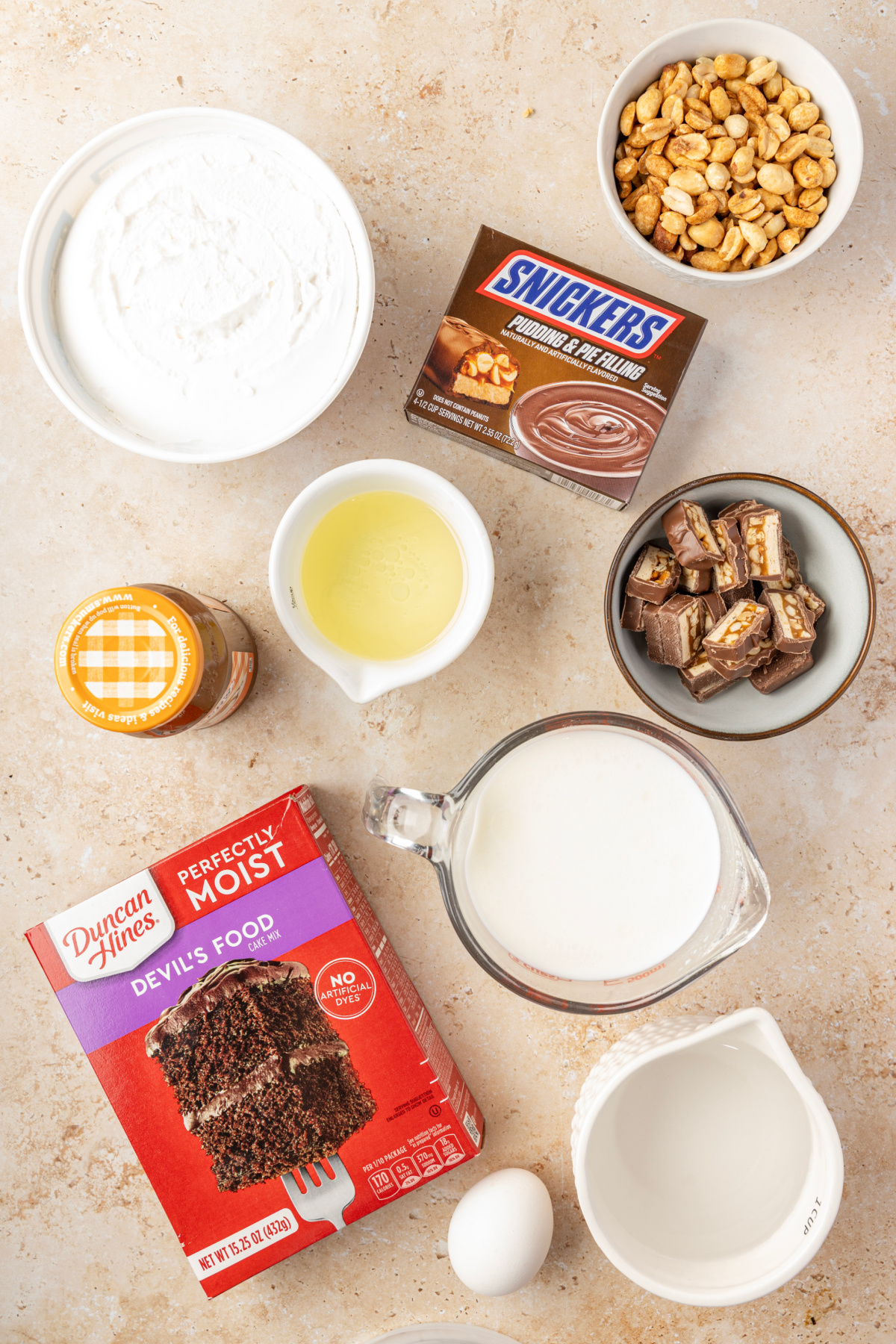 Chocolate cake mix, Snickers pudding, milk, caramel, whipped topping, roasted salted peanuts and Snickers for making the poke cake Ingredients for Snickers poke cake with chocolate cake mix, pudding, caramel, peanuts and chopped Snickers bars