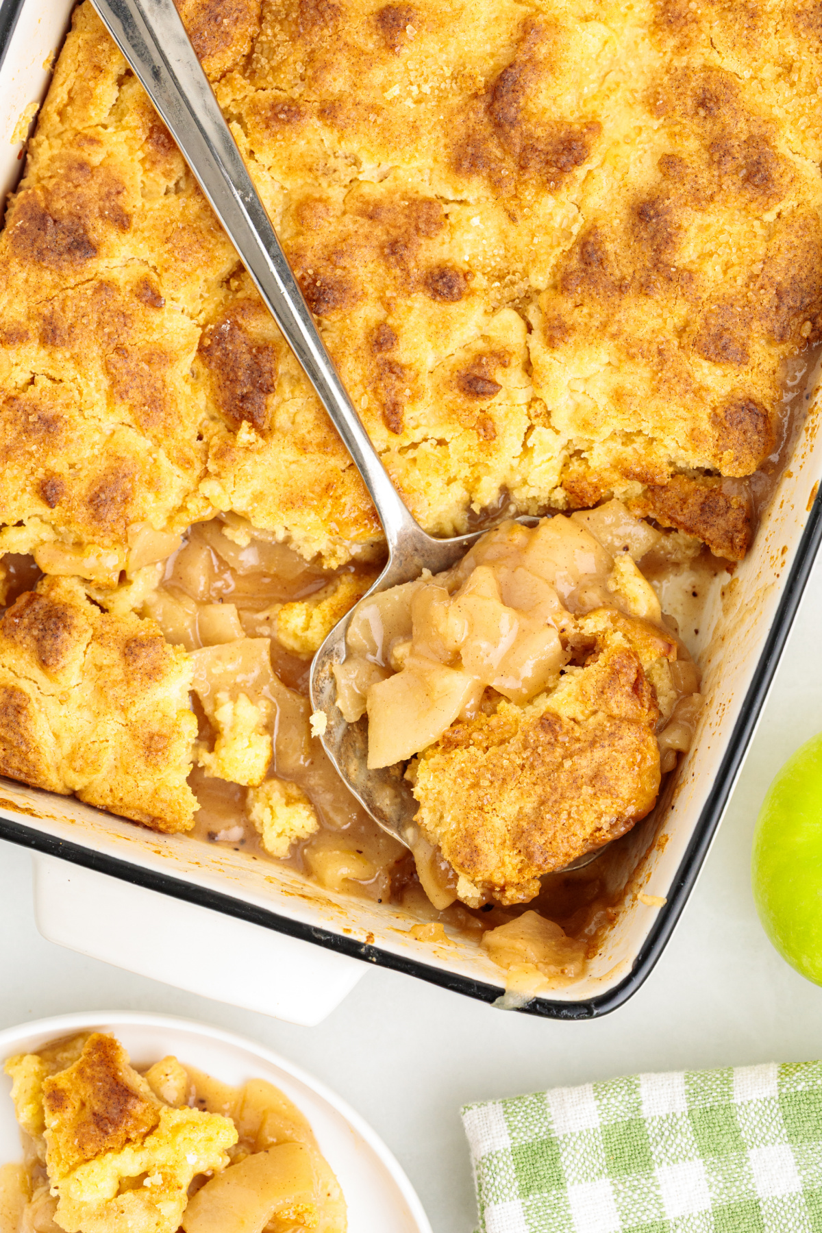 Warm apple cake cobbler with tender Granny Smith apples under a golden butter cake topping. Apple cake cobbler with cinnamon-spiced apples and soft butter cake topping being scooped from a baking dish.