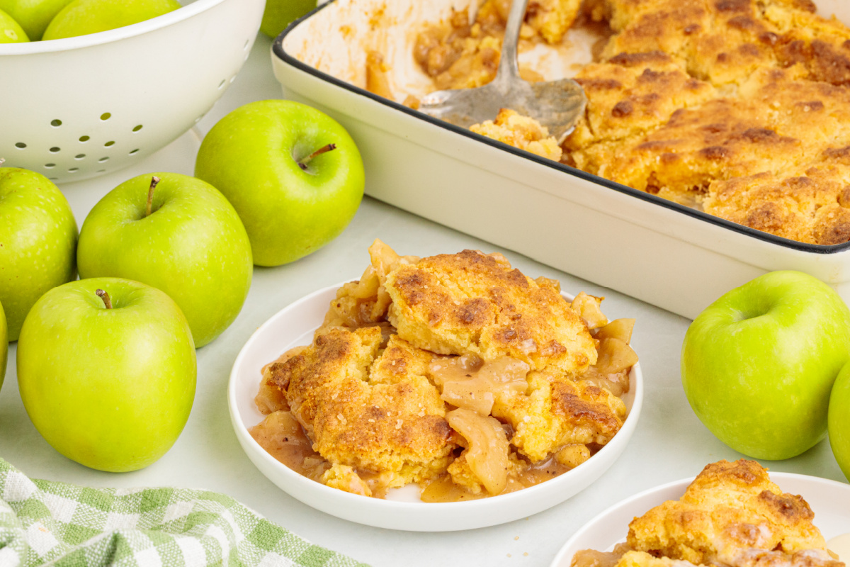 Spoonfuls of warm cinnamon apple filling and soft butter cake served beside the baking dish. Apple cake cobbler served on a plate with saucy Granny Smith apples and pieces of golden cake topping.